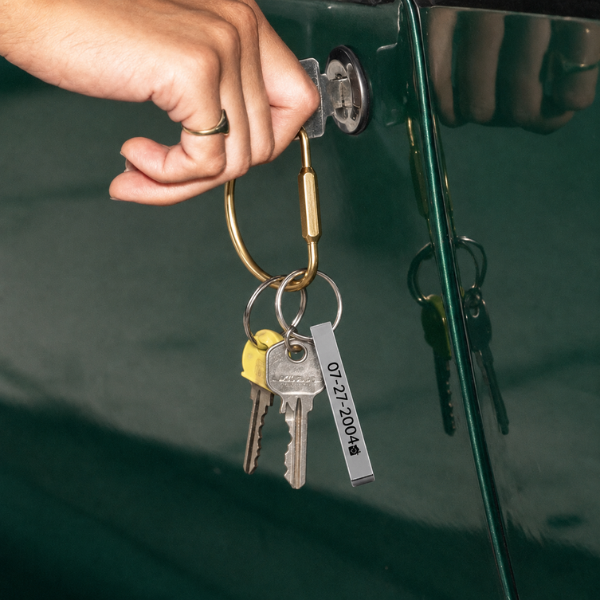 Hand inserting keys into a vintage green car door. Keys have a brass carabiner clip and a rectangular engraved bar keyring with a date. Masculine, lifestyle, editorial feel.